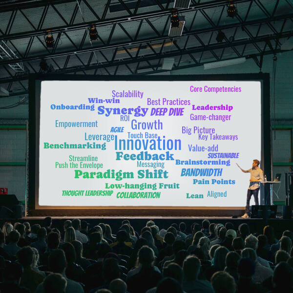 A presenter in front of a large audience at a conference. The screen is displaying a large word cloud with words such as Innovation, Feedback, Growth.